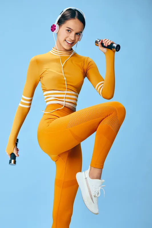 A woman in bright orange athletic wear energizes a dynamic workout with dumbbells, wearing headphones, lifting one knee for balance against a blue backdrop, showcasing fitness, sport, and active