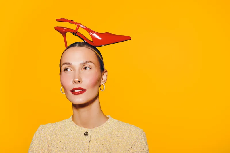 Young woman with red high heel shoe balanced on her head looks away on bright yellow background. Concept for fashion, creativity, and playful style with vibrant colors and modern mood.
