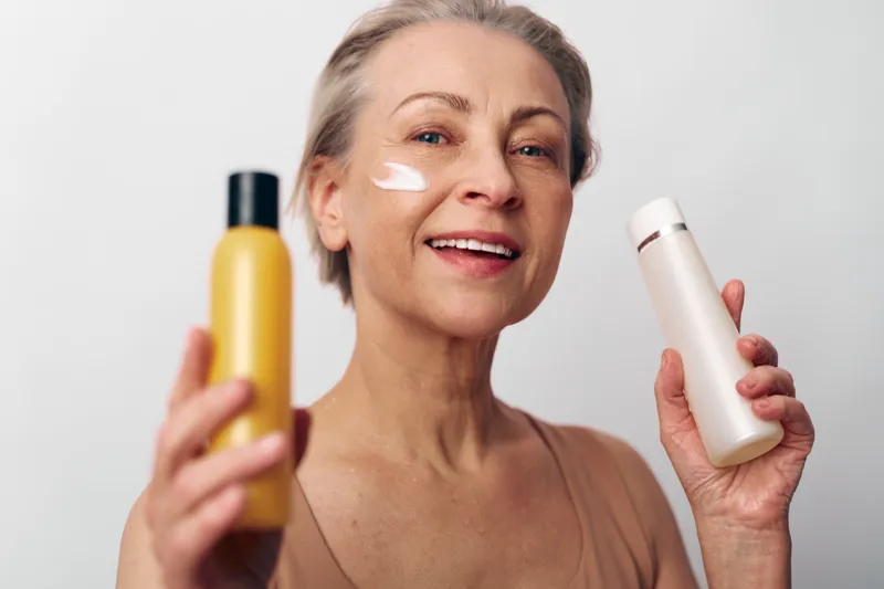 Mature woman with short hair holds two cosmetic bottles smiling, with cream applied on cheek. Concept of skincare, aging beauty, self-care, wellness, and natural skin routine.