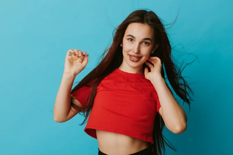 Young woman in red crop top posing and smiling with hand near face against blue background. Casual style, long hair movement, beauty and cheerful expression in studio portrait.