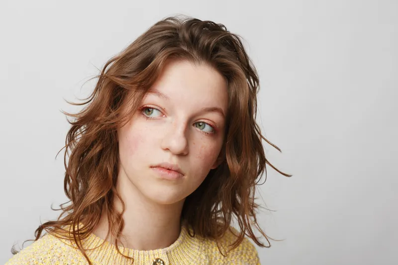 Young woman with curly brown hair and freckles looks thoughtfully to the side against a plain white background. Concept of contemplation, natural beauty, and casual portrait.