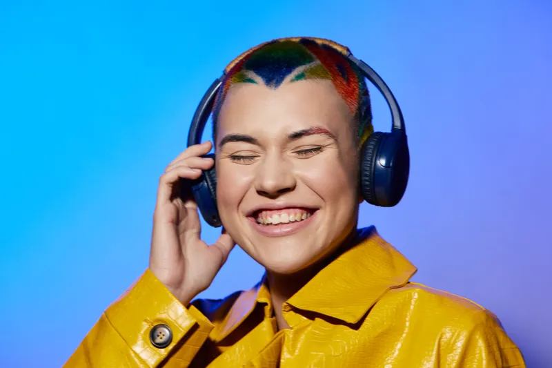 Smiling woman with colorful short hair wearing headphones and a yellow raincoat, enjoying music against a vibrant blue-purple gradient background.