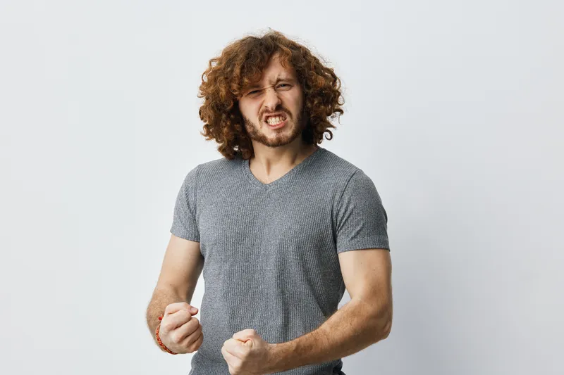 Young man with curly hair expressing anger, wearing a gray t shirt, against a light neutral background, showcasing strong emotions and passion