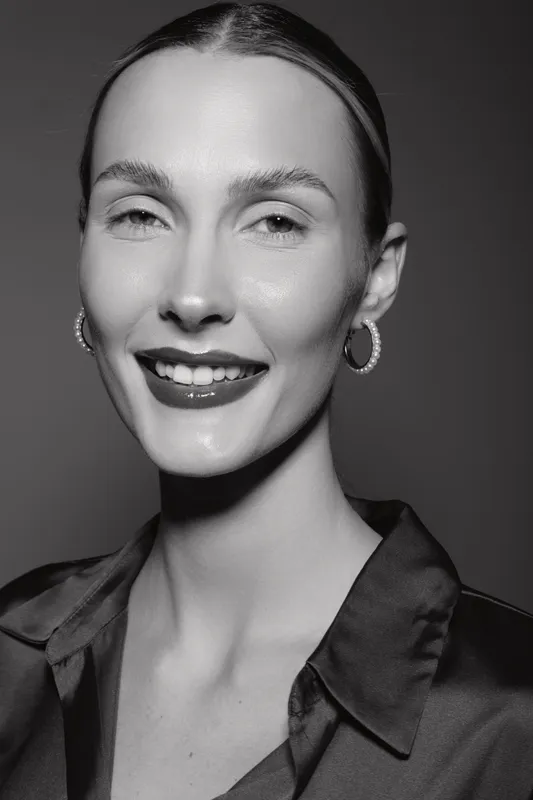 Smiling Caucasian woman with pale skin and slicked back hair wears hoop earrings and dark blouse. Studio portrait in black and white with soft lighting.