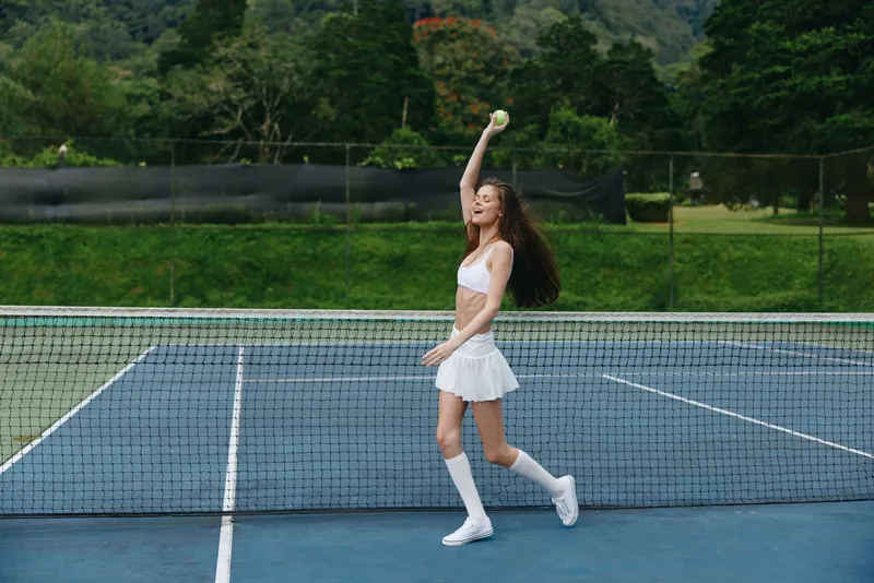 Sporty young woman playing tennis outdoors, showcasing energy and enthusiasm on a sunny day, wearing a white tennis outfit with a scenic green background