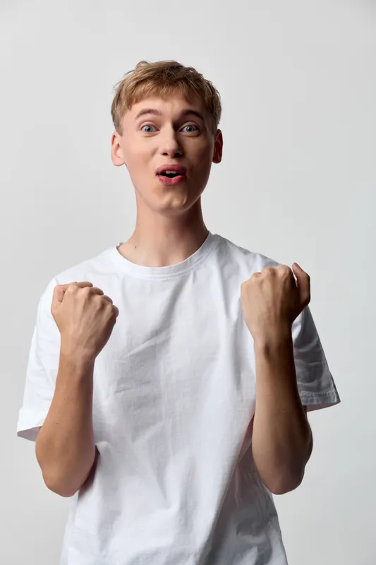 Excited young man in a white t shirt raises fists in celebration, conveying energy, enthusiasm and confidence in a clean studio setting with bright lighting and dynamic pose.