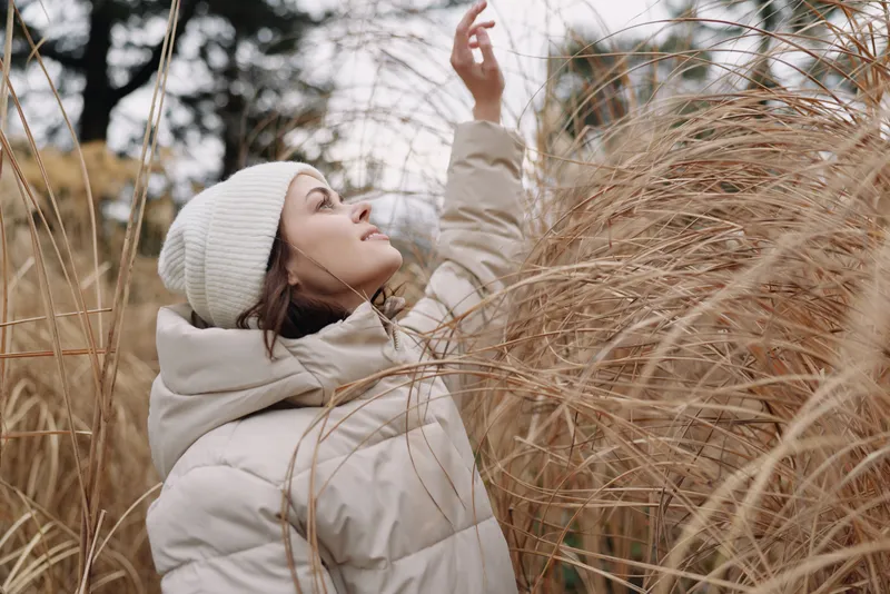 Woman in a warm coat and beanie explores tall grass in a quiet field, capturing a serene moment in nature during cool weather outdoors