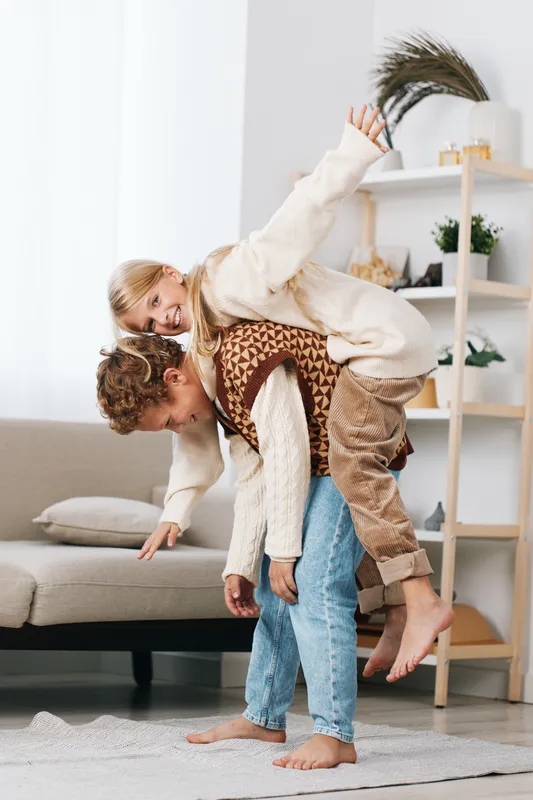 Playful moment of a couple sharing a piggyback ride indoors on a bright living room, warm sweaters, soft rug, relaxed casual scene capturing joy and connection.