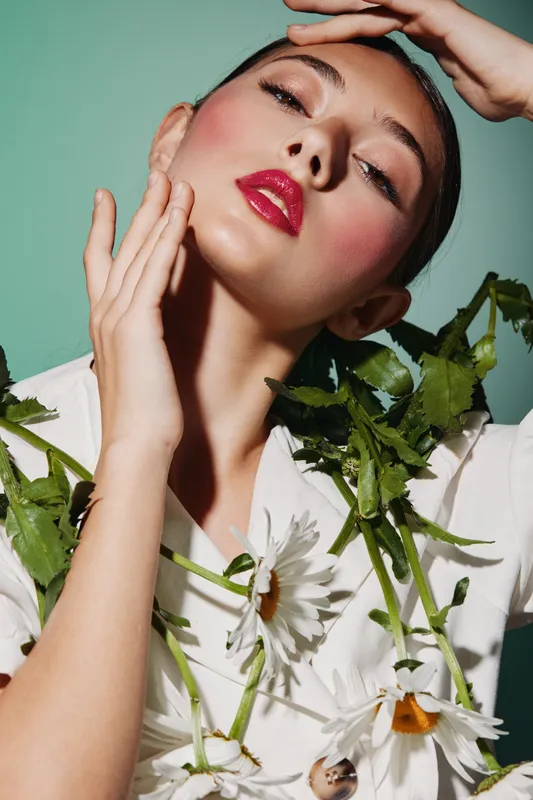 Close-up portrait of a woman with bright makeup and red glossy lips surrounded by white flowers against a pastel background. Concept of beauty, creative fashion and elegance.