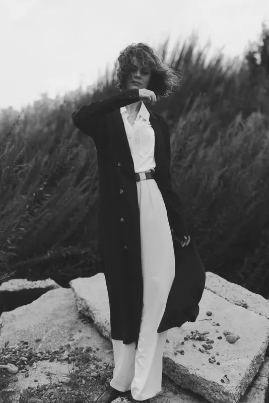 Fashion forward image of a stylish woman in a long coat over white jumpsuit on rocky terrain, wind swept, black and white aesthetic, elegant street style inspiration