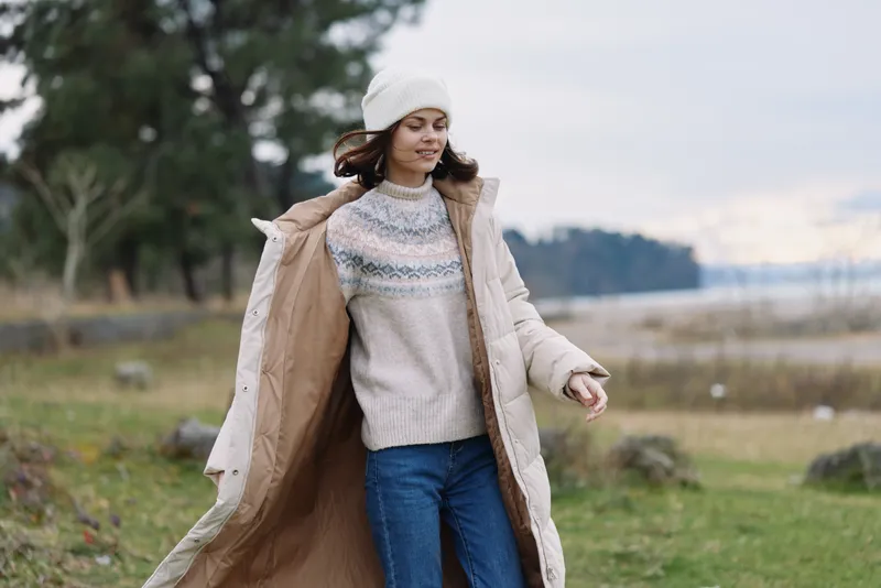 Woman walking outdoors in a long beige coat and knit sweater on a windy day, near a lakeside park with trees and open field in the background.