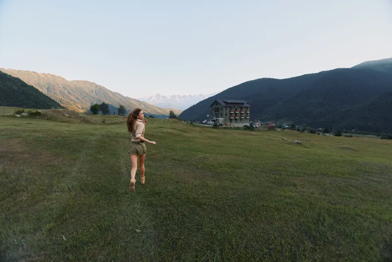 Woman walking in a wide meadow with distant mountains, light breeze, casual dress, enjoying nature, tranquil countryside scenery, open sky, small house in the distance, peaceful outdoor lifestyle