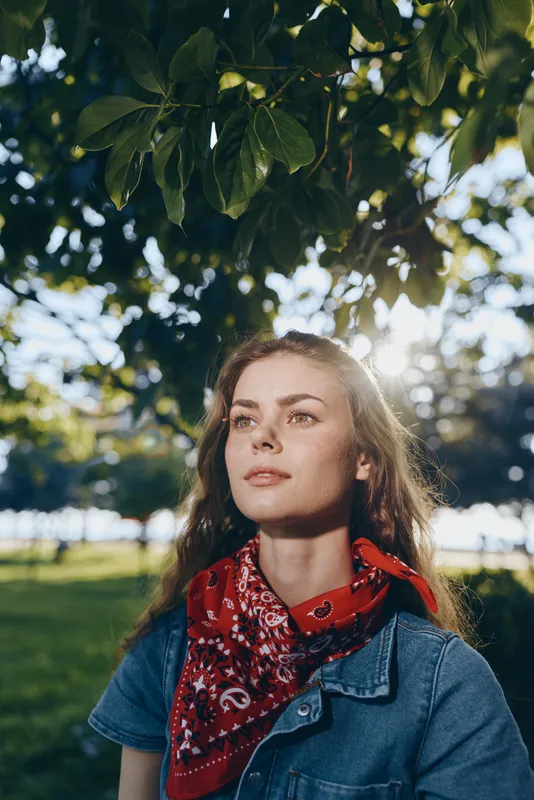 Lifestyle woman in modern boho-western denim street style wearing a red bandana outdoors under green trees with natural sunlight and film color effect.