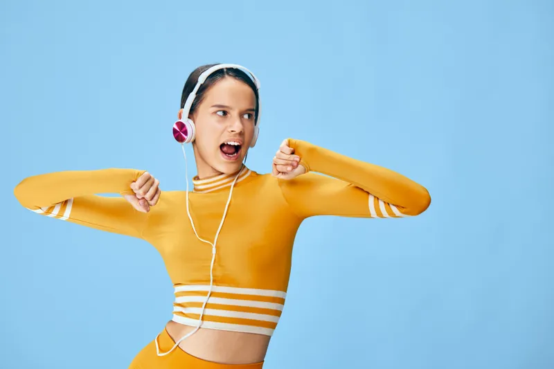 Woman fitness enthusiast dancing in bright orange athletic wear with headphones, posing energetically against a vibrant blue backdrop, showcasing active sport lifestyle and cheerful motion.