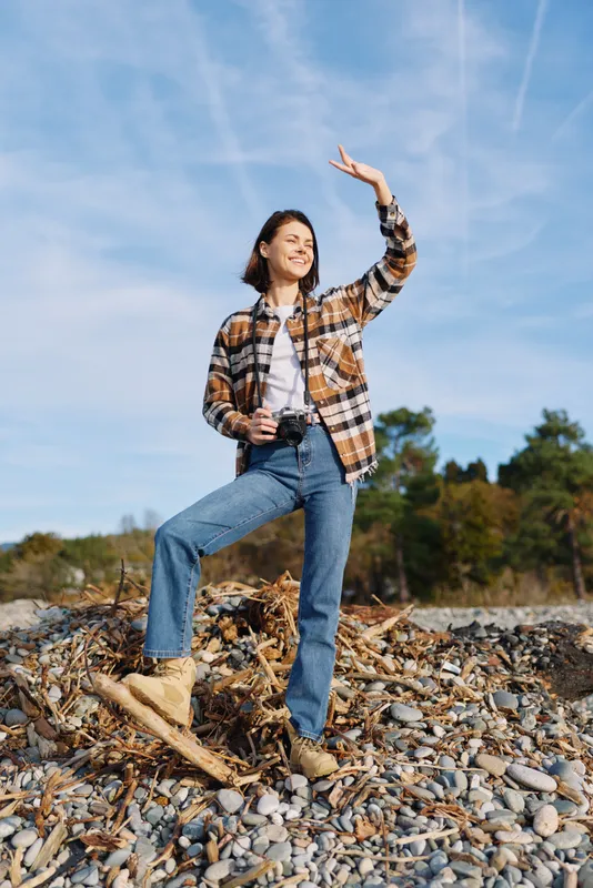 Woman outdoors posing in a casual plaid shirt and jeans, boots on rocky ground under a blue sky, carefree expression and natural light, energetic pose in nature and open space for lifestyle and