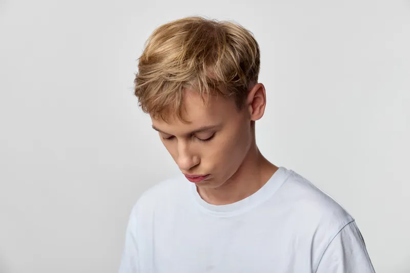 Young man with a downward gaze in a white shirt, captured in a minimalist studio setting. A quiet, contemplative mood highlighting inner focus and everyday simplicity, soft background