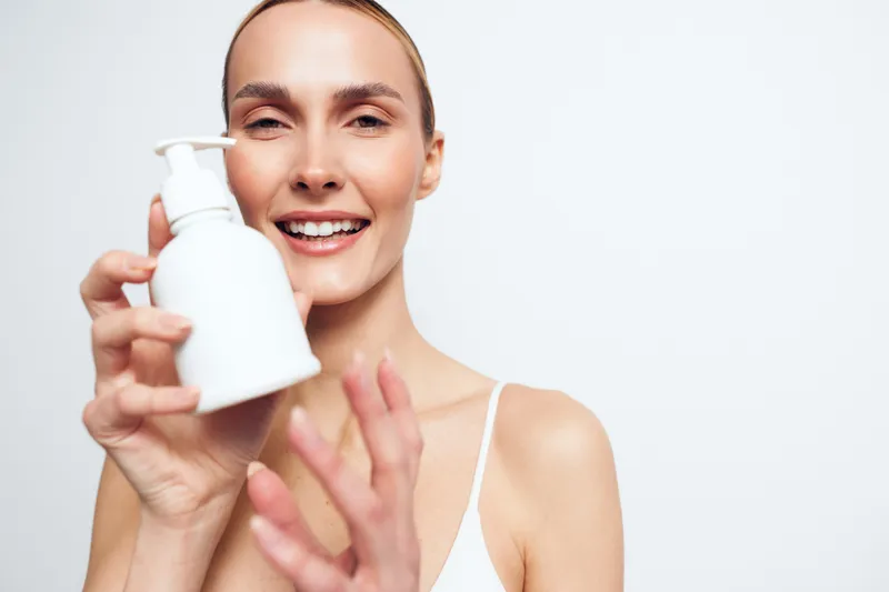 Smiling woman holding white lotion bottle with pump dispenser on white background. Concept of skincare, beauty, wellness, self-care, hydration, and healthy skin. Bright lighting.