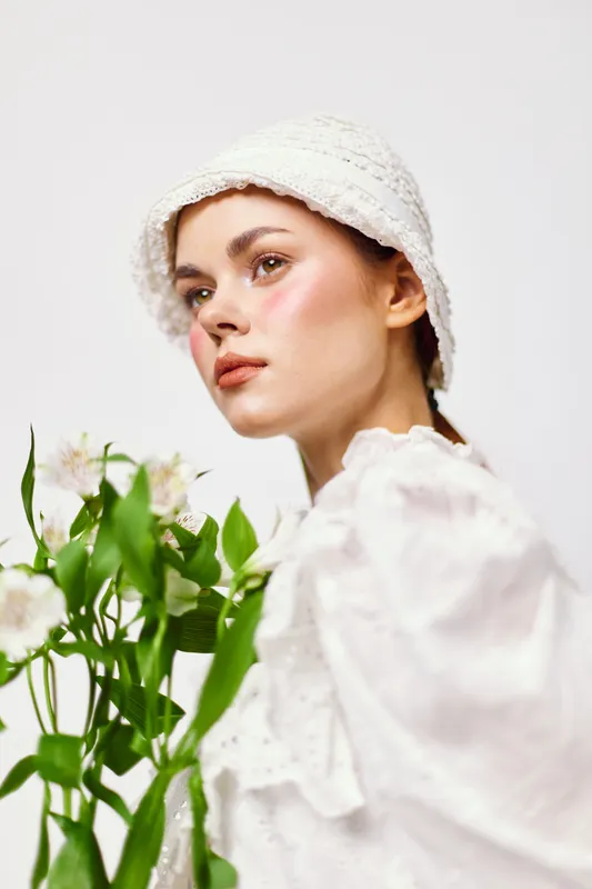 Young woman in a white dress and hat holding flowers, expressing serenity and freshness in a minimalist setting, perfect for seasonal concepts
