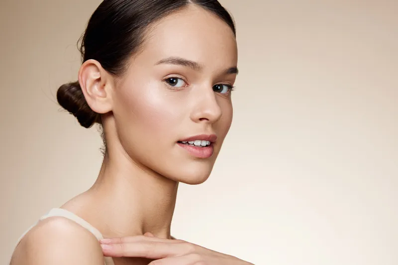Portrait of a young woman with flawless skin, cheerful expression, wearing minimal makeup and a light top, beautiful neutral background, promoting natural beauty and skin health
