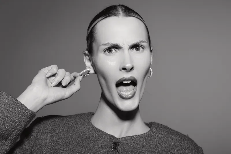 Young white woman with light skin and slicked hair shows frustrated expression and pulls on earring in studio with gray background.