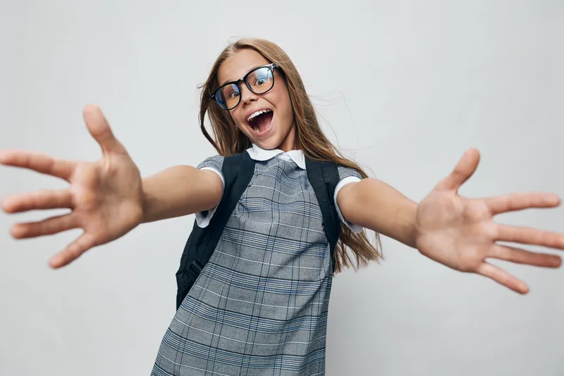 Happy girl with long hair and glasses reaching arms forward smiling for fun and excitement with school dress on gray background in studio interior.