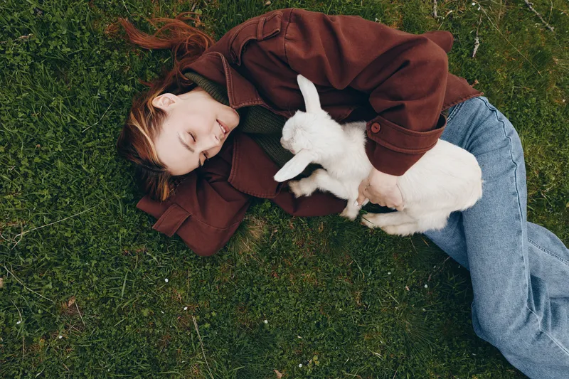 Relaxed lifestyle moment with a young woman lying on grass, cuddling a white goat kid, smiling softly beneath a brown sweater, natural outdoor scene, wholesome bond, candid countryside vibe, peaceful