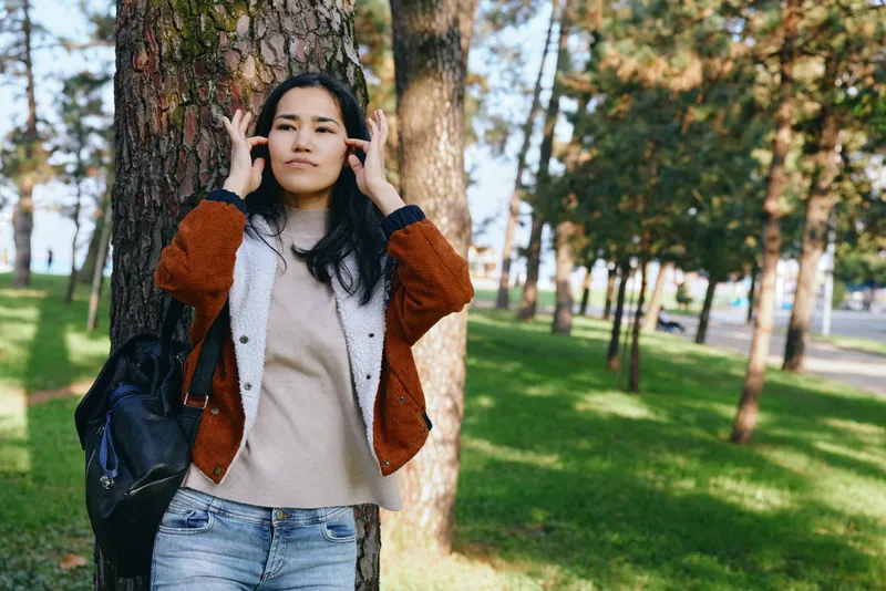 Outdoor scene with a woman by a backpack in a leafy park, wearing a warm cardigan and denim jeans, enjoying a sunny day among tall trees in gentle light.