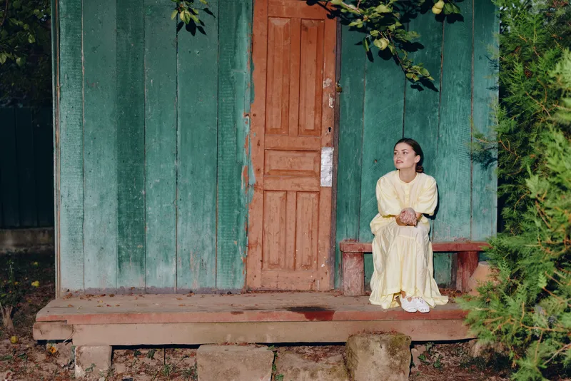 Portrait of a woman wearing a pale yellow dress sitting on a wooden porch beside a weathered green house in a quiet outdoor setting