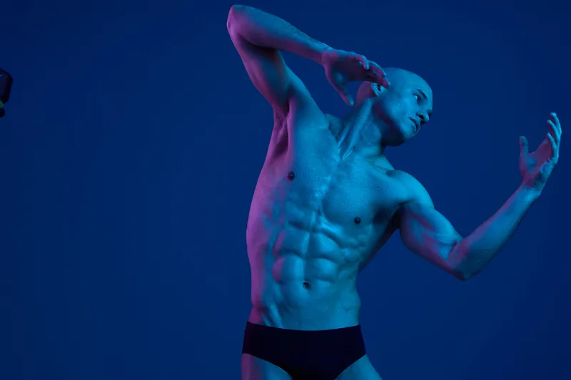 Athletic man posing dramatically under moody blue lighting, showcasing muscular physique and expressive gestures in a captivating artistic moment.