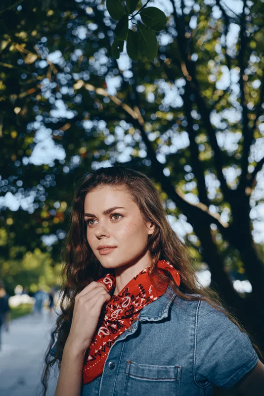 Young woman with curly hair wearing denim shirt and red bandana outdoors with natural sunlight and leafy background in warm summer atmosphere.