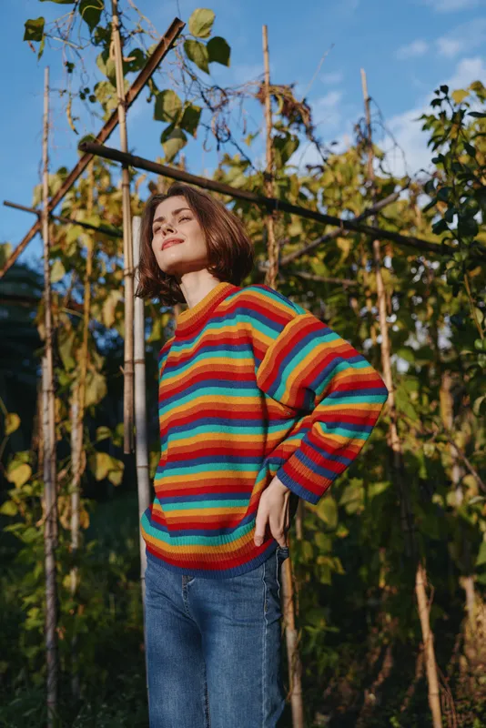 Woman in a colorful striped sweater outdoor in a garden, smiling young adult in casual outfit and denim jeans, short curly hair, standing confidently in sunlight for lifestyle fashion.