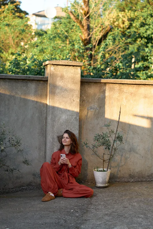 Cheerful woman outdoors, seated on the ground, red outfit, phone in hand, sunlit moment, relaxed posture, warm tones, leafy background, stone wall