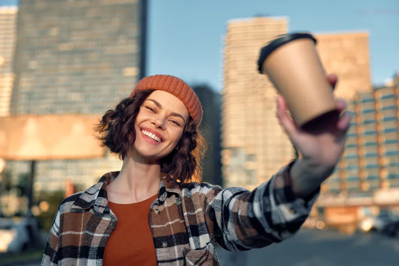 Woman smile holding coffee cup in city during golden hour, candid urban lifestyle portrait with beanie and warm sunlight, authenticity, mindful living and emotional storytelling moment.
