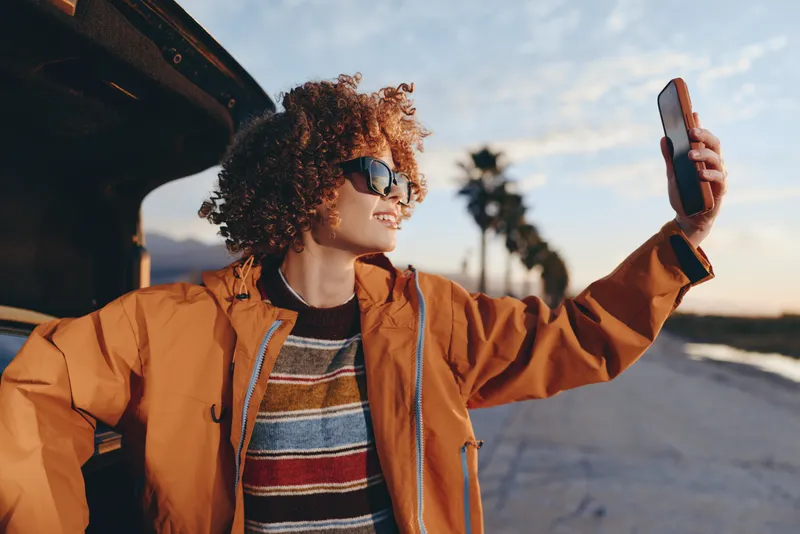 Woman lifestyle outdoors, smartphone in hand, sunglasses on face, curly hair, orange jacket, striped sweater, standing by road at sunset, enjoying communication and travel, lifestyle portrait