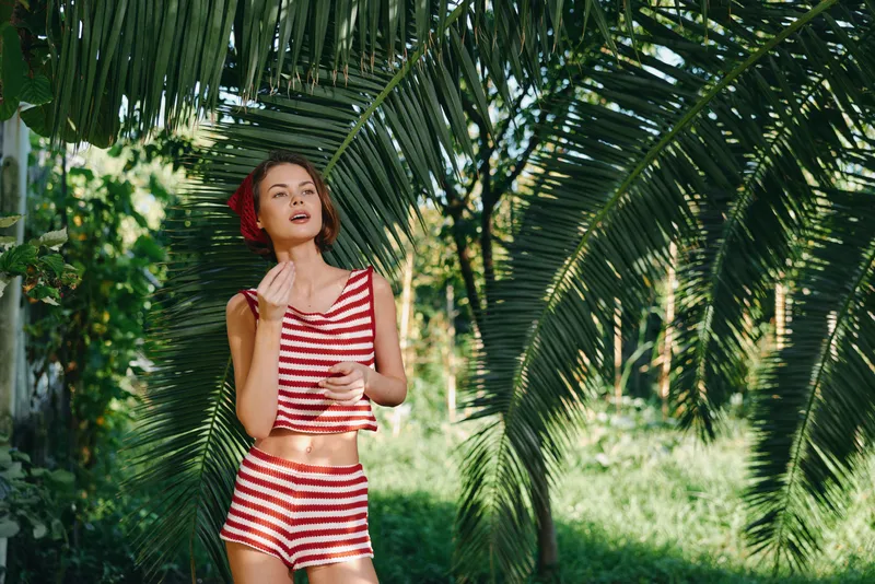 A stylish woman in a red and white striped bikini stands beneath lush tropical palm fronds, basking in warm sunlight, creating a bright outdoor lifestyle moment for fashion and travel stock.