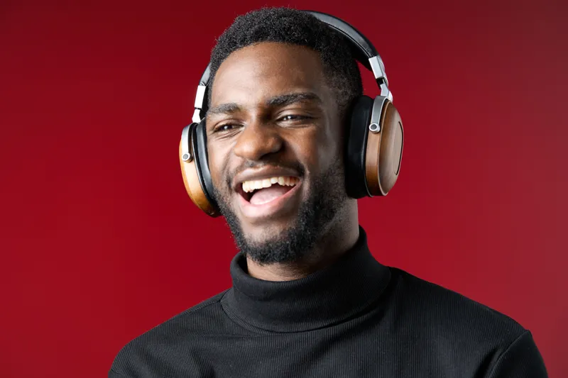 Young man with headphones smiling joyfully against a red background, showcasing music enjoyment and stylish fashion