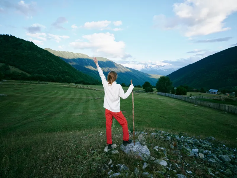 Outdoor landscape with a woman in white top and red pants standing on a rocky hill, arms raised toward the vast valley and mountains, celebrating nature, freedom and adventure
