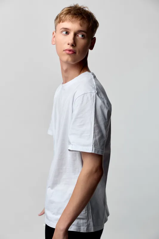 Young man wearing a white T shirt poses in a studio with a subtle side glance, showcasing casual fashion, simple backdrop, and modern lifestyle appeal for stock use.