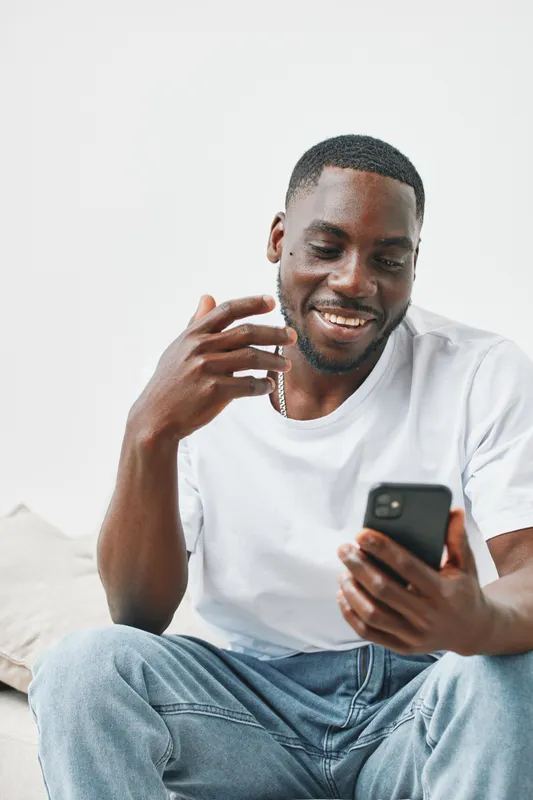 Smiling African man in casual white t shirt, enjoying video call on smartphone, relaxed home environment with neutral background
