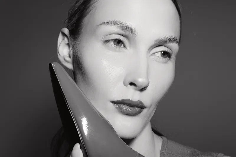 Close-up portrait of a caucasian woman with fair skin holding a shiny high heel shoe near her face. Concept for fashion, elegance, beauty, and style in monochrome.