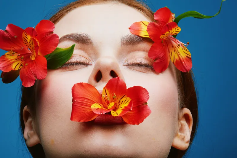 Woman with closed eyes adorned with vibrant red flowers on a blue background, showcasing natural beauty and serenity