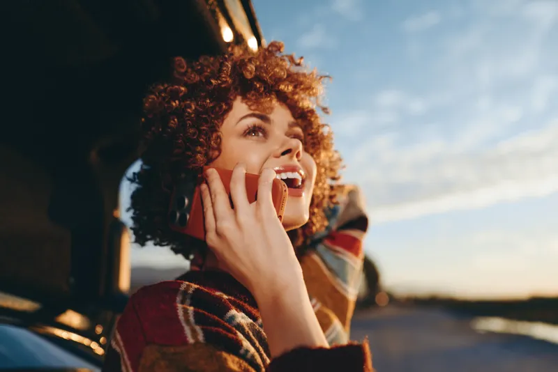 Woman lifestyle curly hair smiling outdoor sunset plaid coat snack in hand, warm evening light, candid travel moment, relaxed posture, soft bokeh, natural colors