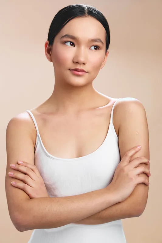 Asian woman stands with arms crossed in a neutral studio setting, her calm expression and soft lighting convey confidence and focus.