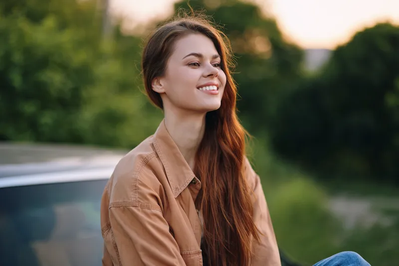 Young woman in a casual outfit smiling outdoors, reflecting joy amidst greenery in a warm setting, representing a carefree lifestyle and connection with nature.