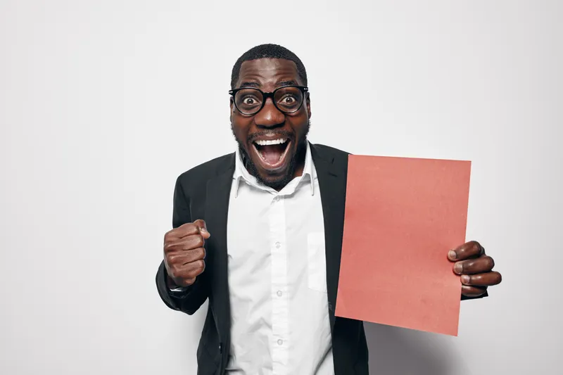 African American man with dark skin wearing glasses and suit holding red paper with excited expression and clenched fist on white background for success and achievement.