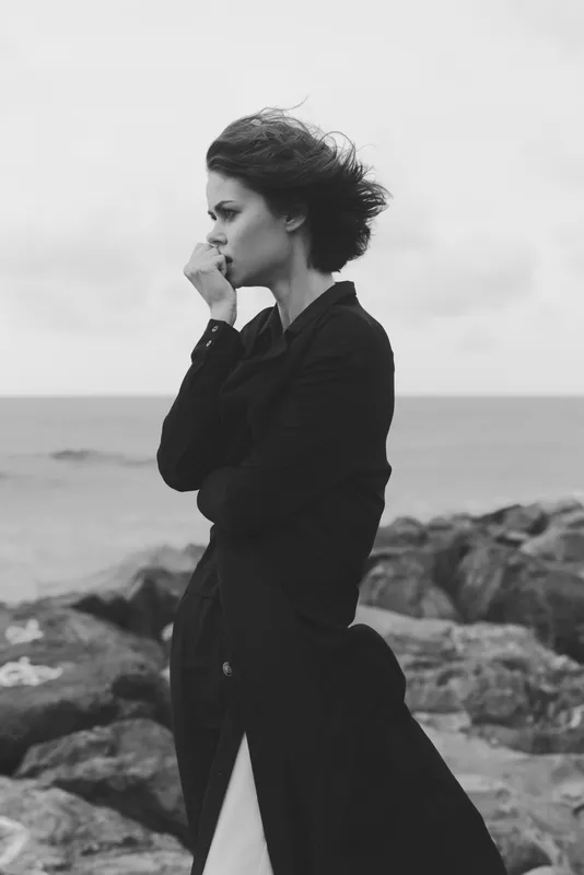 Woman stands on a rocky coastline, sea in view, wind blowing, wearing a long coat in a moody black and white outdoor scene, fashion and solitude in a dramatic seascape