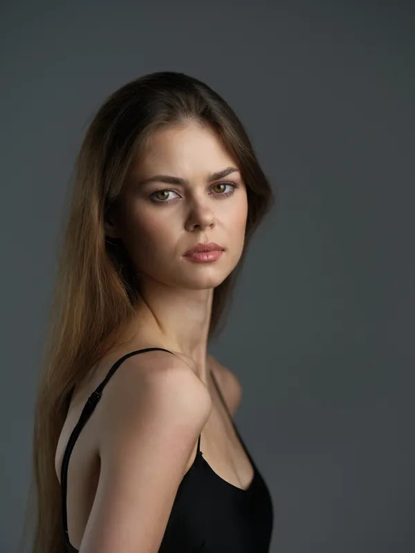 serious woman with long hair posing against gray background in black top, natural light, portrait, soft mood