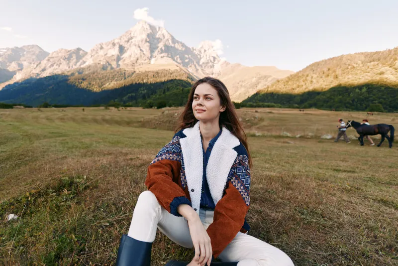 Woman portrait sitting in meadow with mountain backdrop and horse in distance, outdoors in countryside nature during golden hour. Cozy jacket, relaxed pose and wide valley landscape.