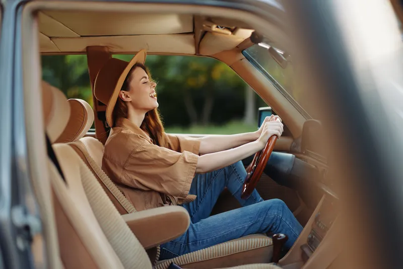 Young woman wearing a stylish hat and casual outfit joyfully driving a vintage car on a sunny day, embodying freedom and adventure.
