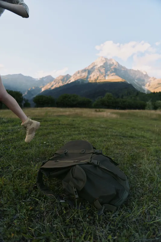 A hiking woman runs across a grassy field with a backpack on the ground, mountains rising in the background, conveying outdoor adventure and focus.